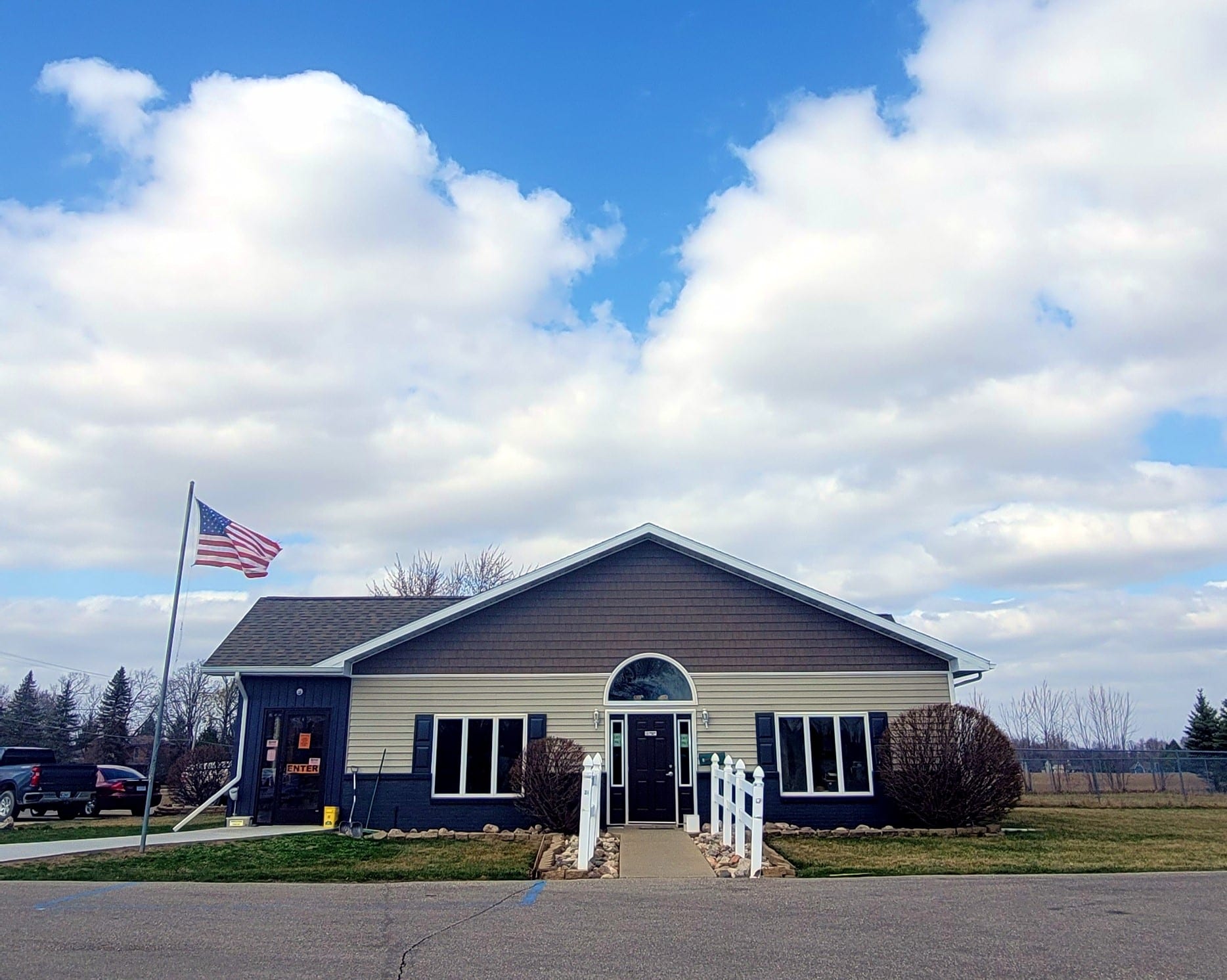 A one-story building with an American flag on the left, under a partly cloudy sky. A path leads to the entrance, flanked by white fences and bushes.