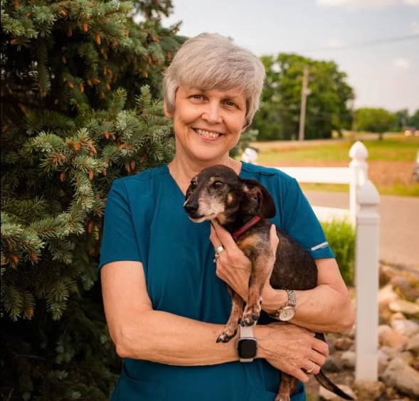 A person with short gray hair, wearing a blue shirt, smiles while holding a small brown and black dog outdoors. Trees and a fence appear in the background.