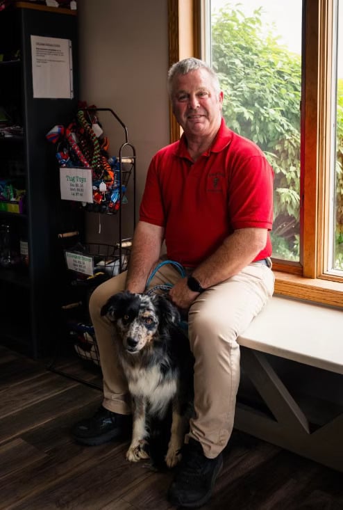 A man in a red shirt sits by a window, holding a black and white dog, in a room with a wooden floor.