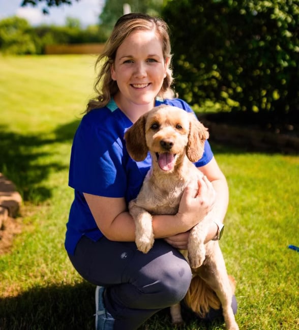 Person kneeling on grass, smiling and holding a brown dog. Both appear relaxed and outdoors, with bright sunlight and greenery in the background.