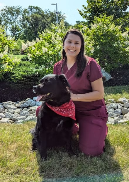A woman kneels beside a black dog wearing a red bandana in a garden setting with rocks and greenery.