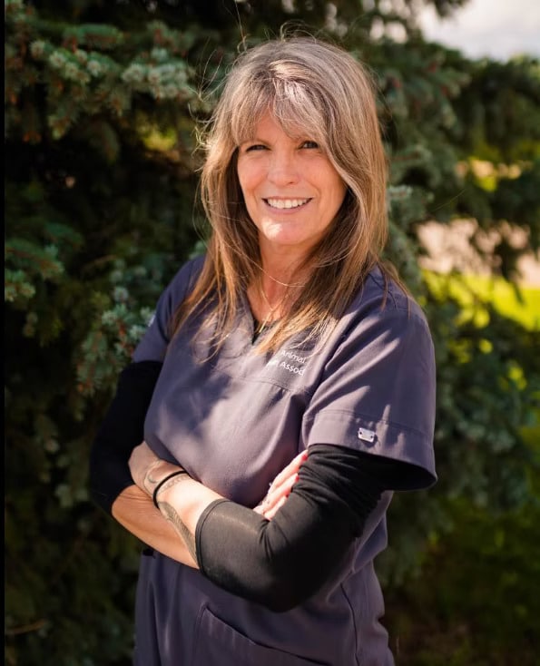 Woman with long hair smiles while standing outdoors in front of a tree, wearing a gray medical scrub top and arms crossed.