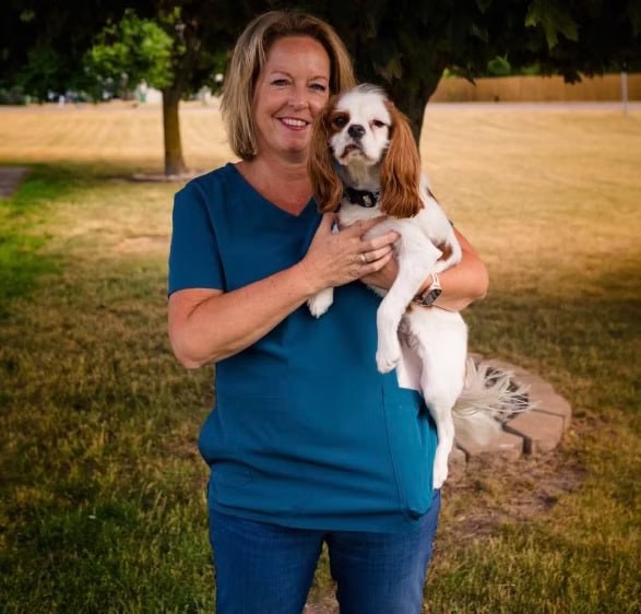 Woman in a teal shirt holding a small white and brown dog, standing on grass with trees in the background.