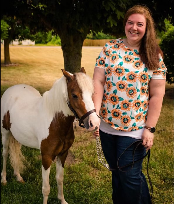 A woman in a floral shirt stands on grass next to a small white and brown pony, holding its leash.