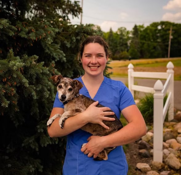 Person in blue scrubs smiling and holding a small dog outside near a tree and a white fence.
